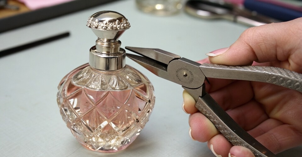 A close-up shot of pliers carefully gripping the edge of a crimped metal collar on a perfume bottle, bending it upwards.